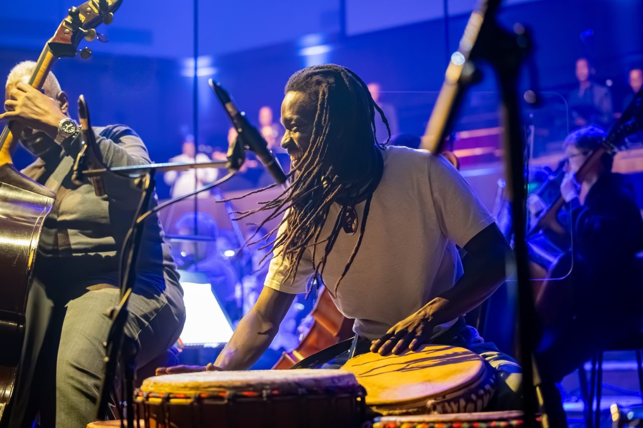 A smiling man looks to the side while playing African percussion.