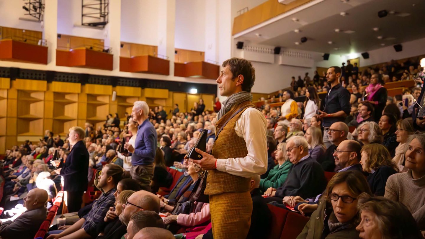 Audience members holding metronomes stand in busy concert hall.