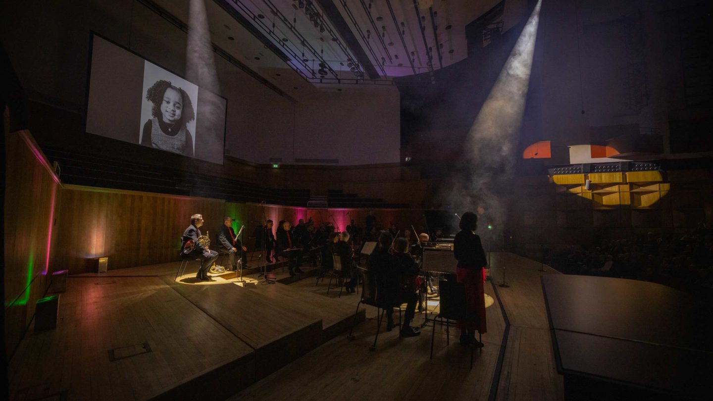 Dark concert hall stage, with projected image of young girl on a screen, and individual musicians highlighted by spotlights.