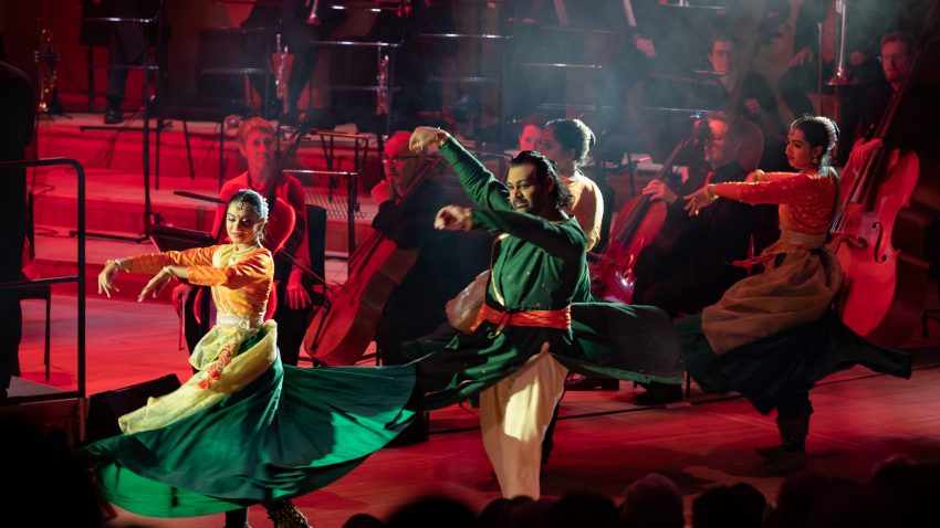 Dramatically lit traditional south Asian dancers perform, with orchestral players dimly lit in the background.