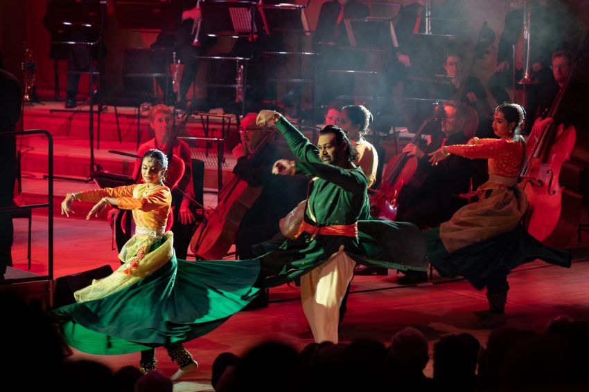 Dramatically lit traditional south Asian dancers perform, with orchestral players dimly lit in the background.