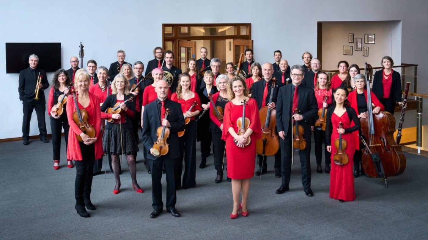 Musicians wearing red and black smile and hold their instruments in a foyer space.