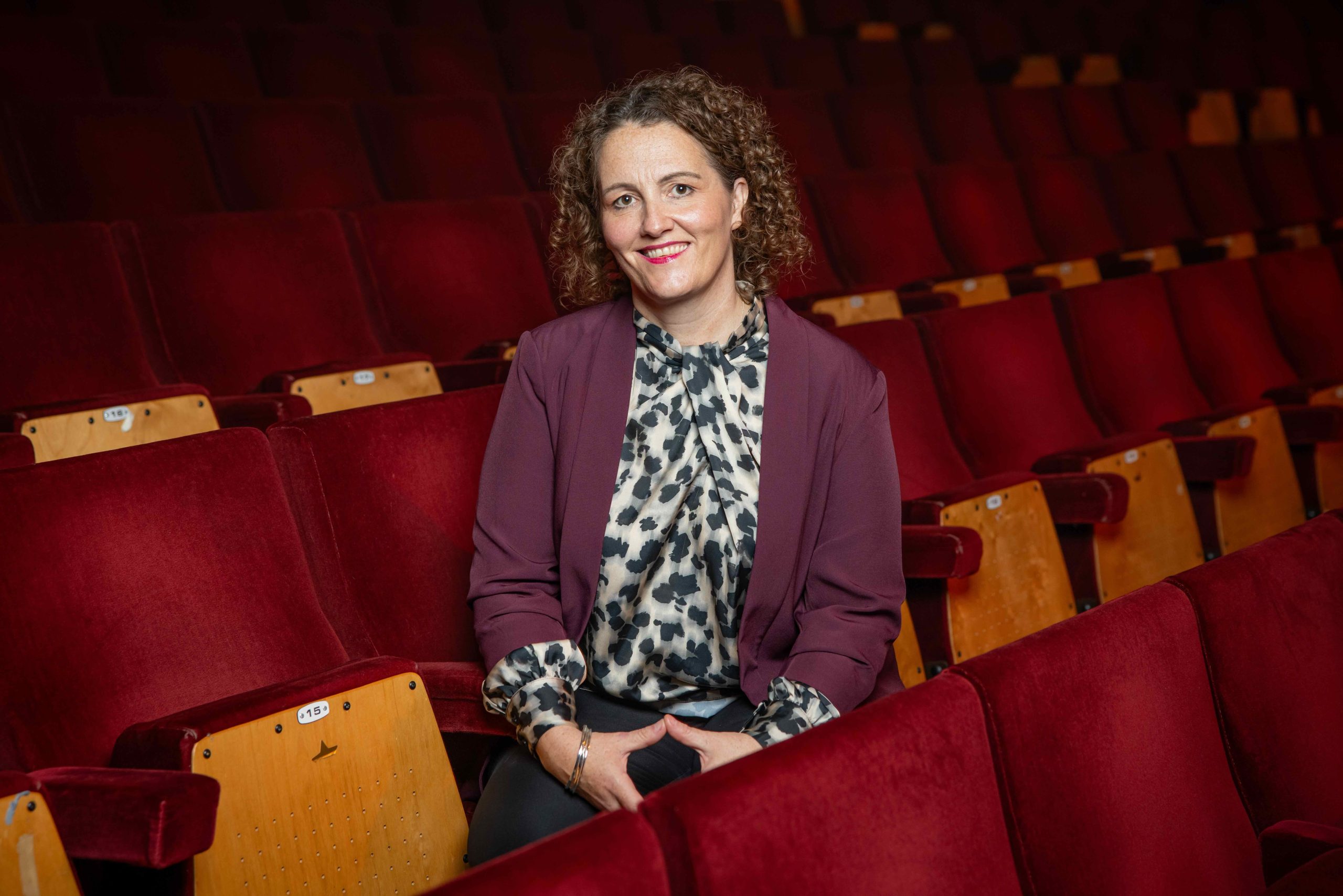 A smiling woman photographed in the red seats of a concert hall.