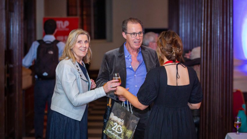 A woman is handed a glass of wine as she looks at the camera.