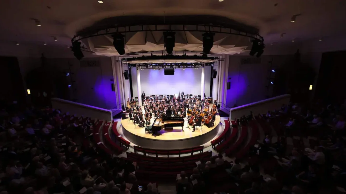 A tightly packed orchestra on a circular stage with a piano in a dimly lit concert hall.