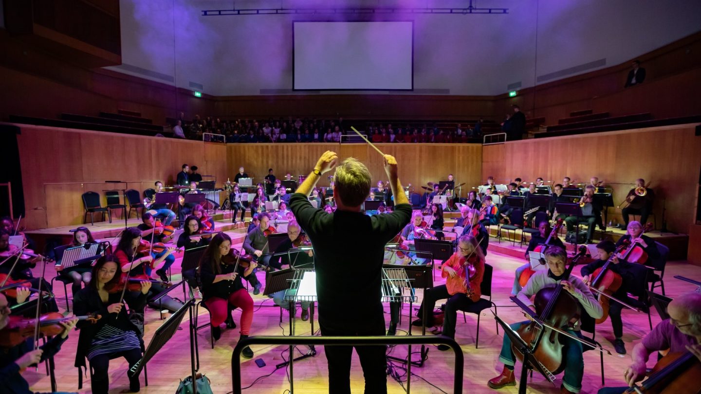 Dramatic photo of a conductor standing on stage in front of musicians and a choir.