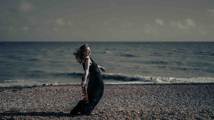 Woman with a violin in one hand leans backwards on a pebble beach.