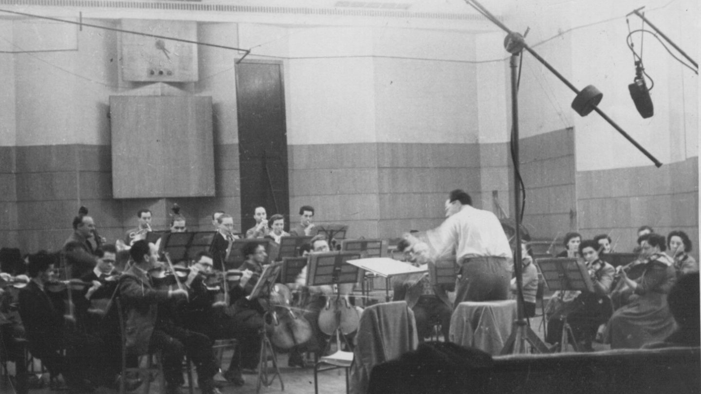 Old black and white photo of orchestra and conductor in a large studio. Microphones hang above them.