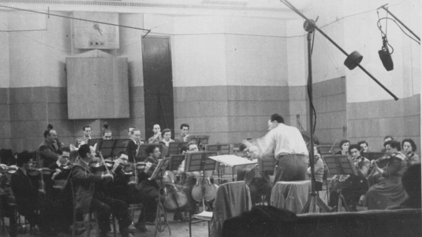 Old black and white photo of orchestra and conductor in a large studio. Microphones hang above them.