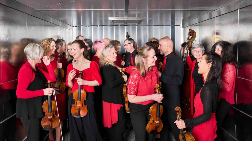 Musicians in black and red gather inside a large lift with their instruments.