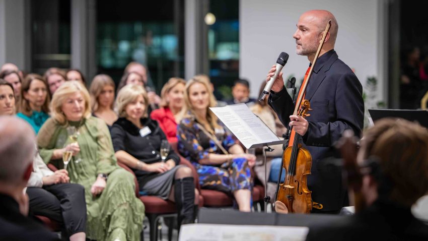Man holding violin addresses audience with microphone.
