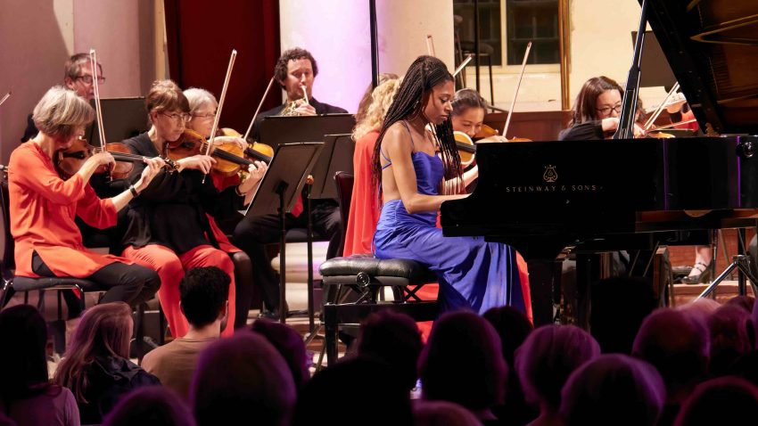 Woman plays piano alongside orchestra.