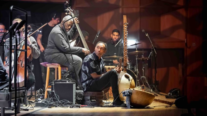 A man sits playing the kora on stage with other musicians behind him.