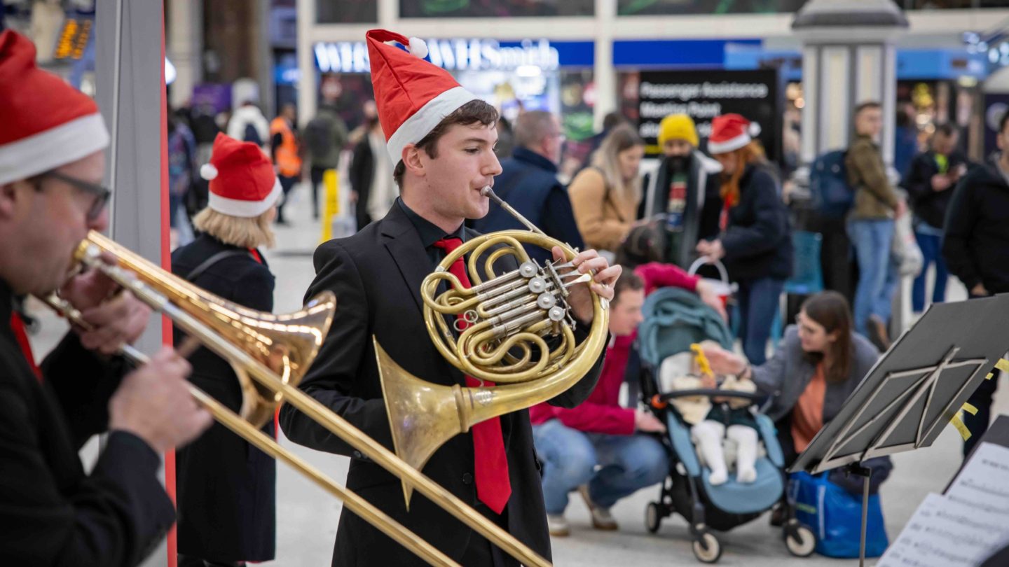 Man in santa hat plays the horn on a busy station concourse.