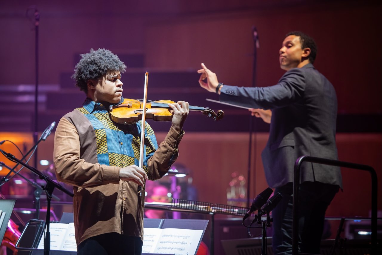 A man shuts his eyes while playing the violin and a conductor raises his hand in the backround.