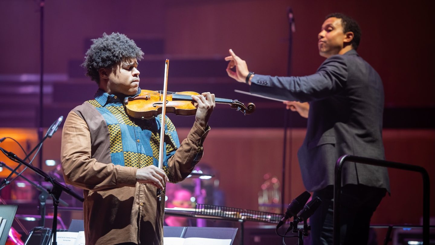 A man shuts his eyes while playing the violin and a conductor raises his hand in the backround.