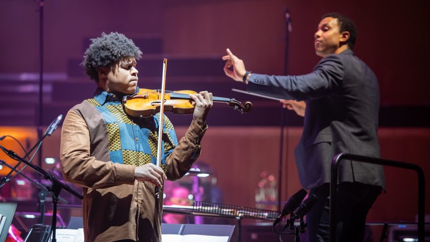 A man shuts his eyes while playing the violin and a conductor raises his hand in the backround.