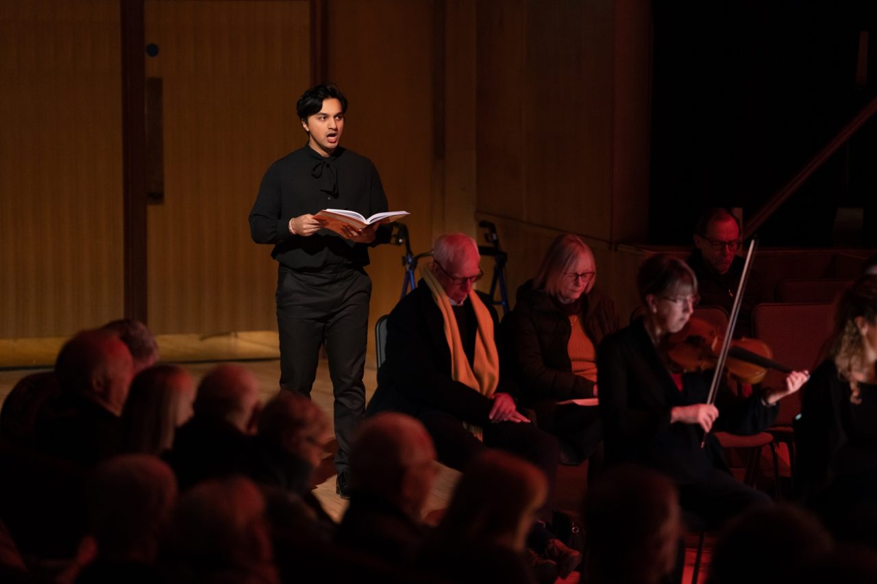 A man dressed in black sings whilst standing behind audience members.