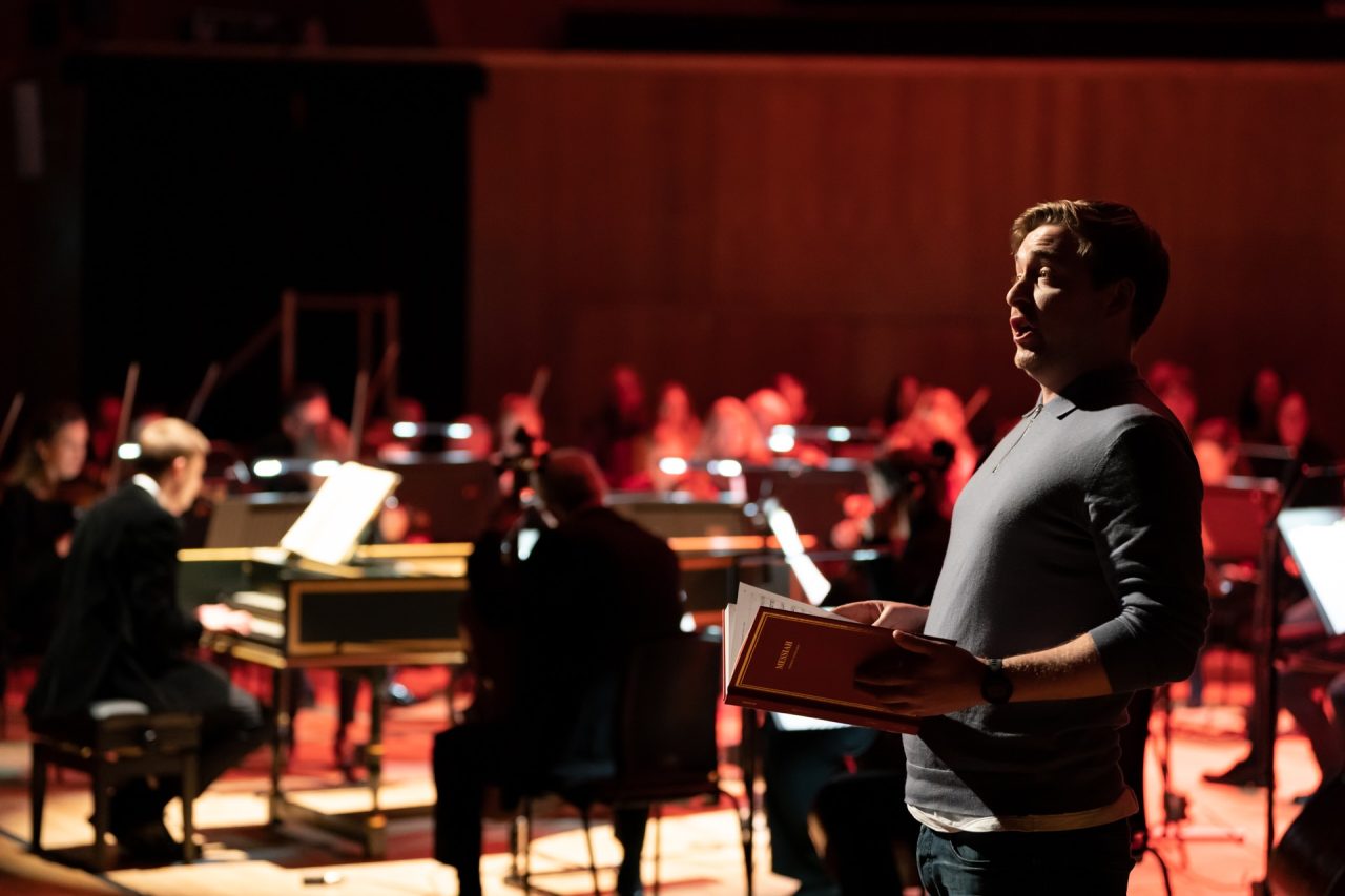 A man in a grey t-shirt leans backward and sings from a book.