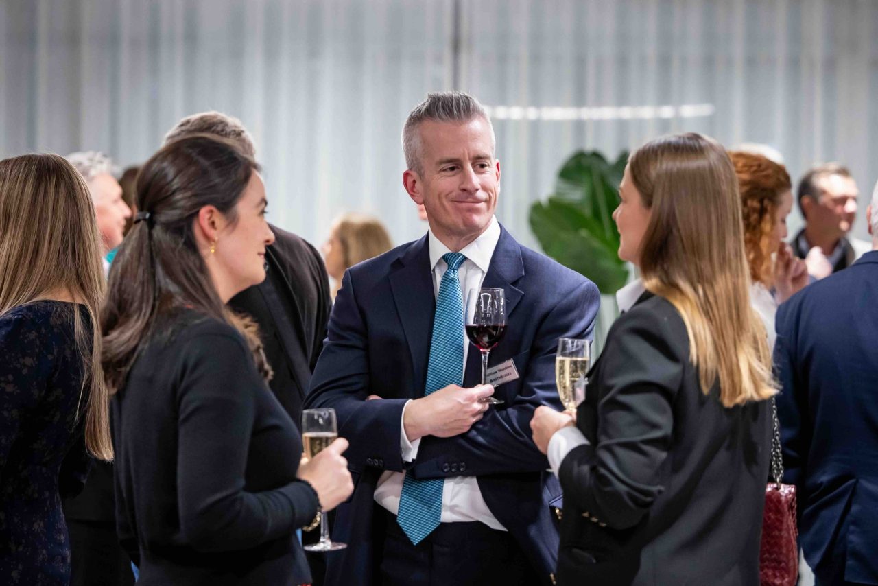 Man in suit holds a glass of wine next to colleagues.