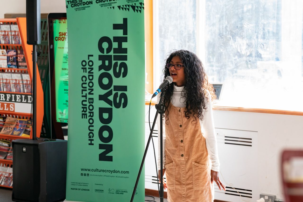 A young girl in a pinafore sings into a microphone.