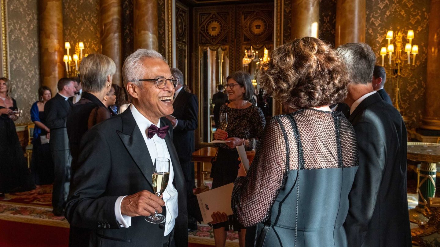 A man in black tie smiles in a grand room, holding a full champagne flute.