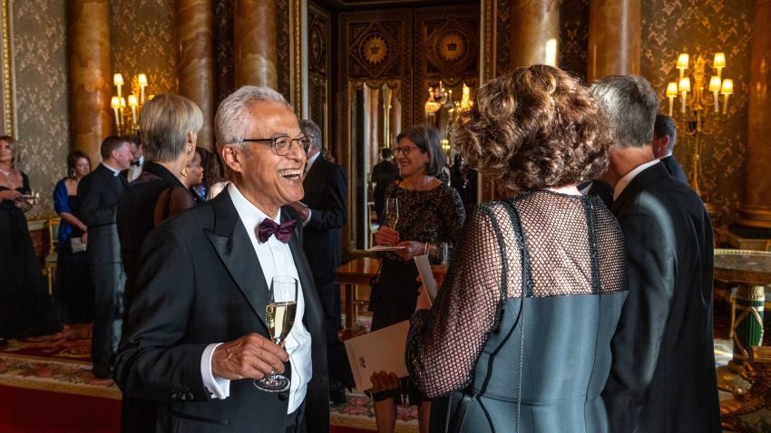 A man in black tie smiles in a grand room, holding a full champagne flute.