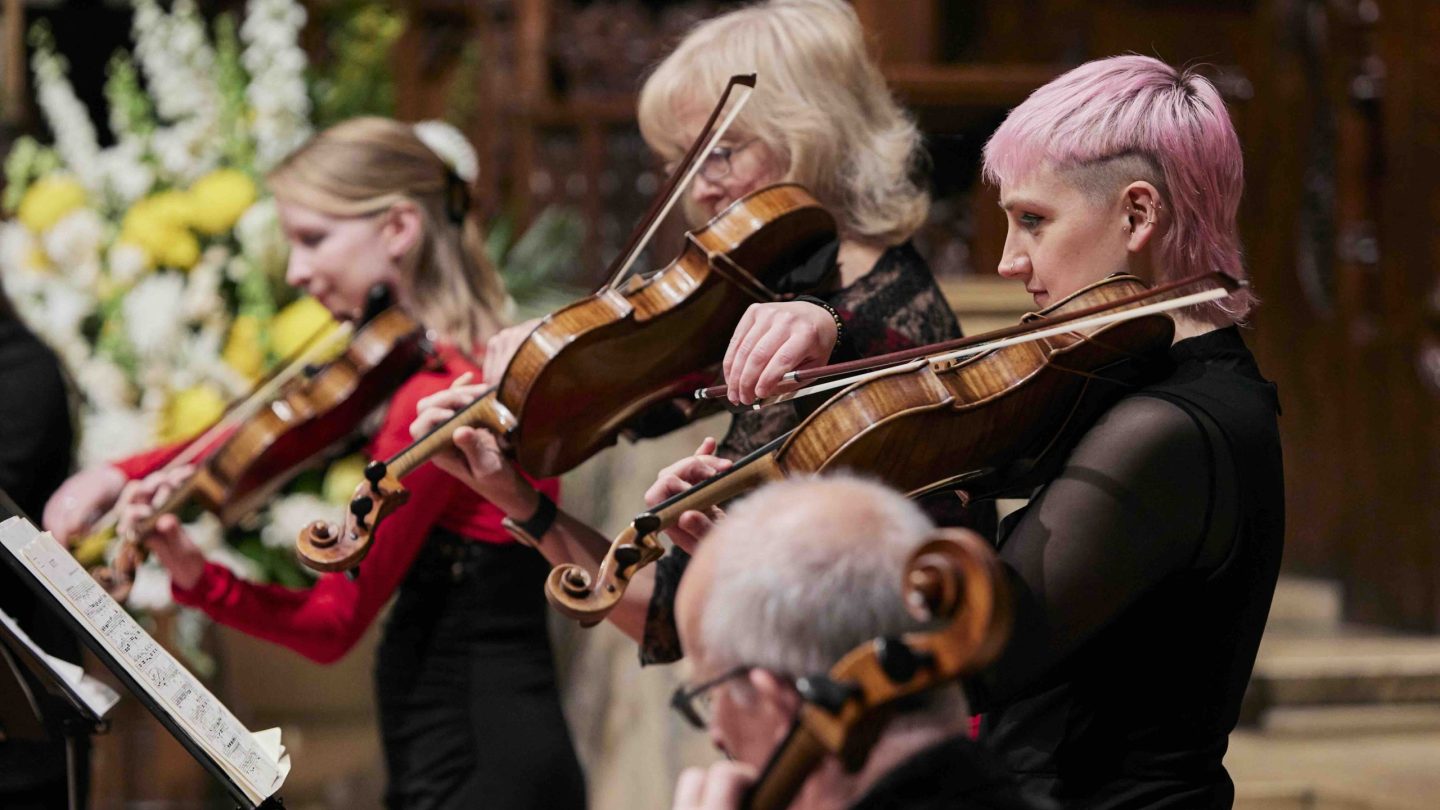 A woman with pink hair looks at music on a stand whilst playing the viola.