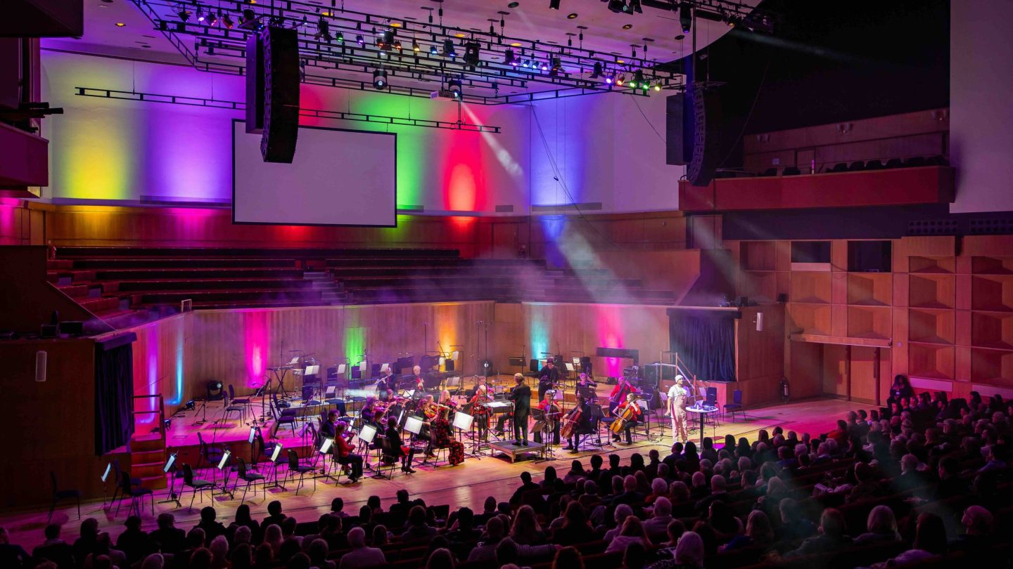 An audience watches an orchestra on stage while colourful rays of light beam down on the players.