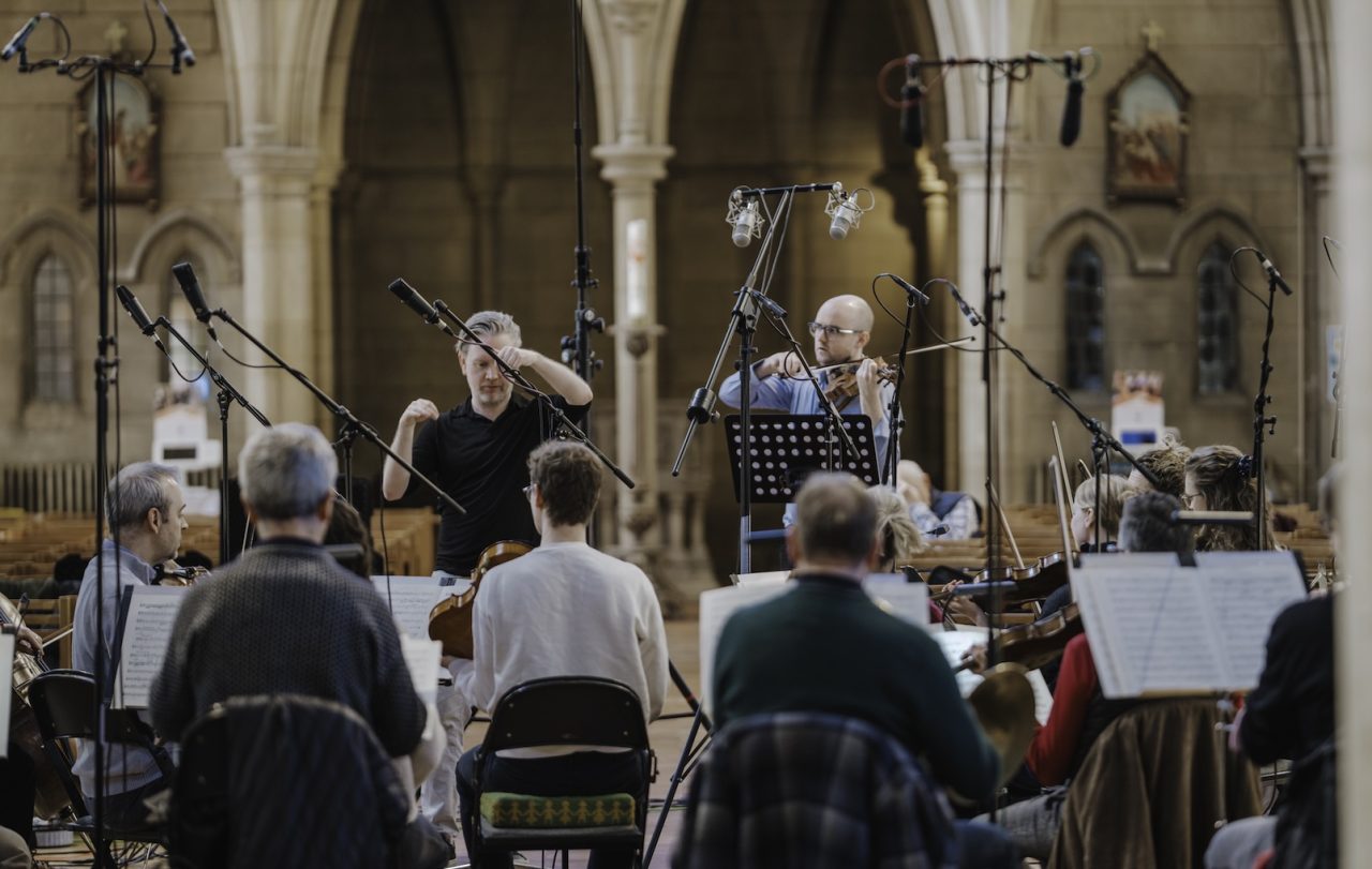 A conductor stands next to a man playing the violin surrounded by microphones.